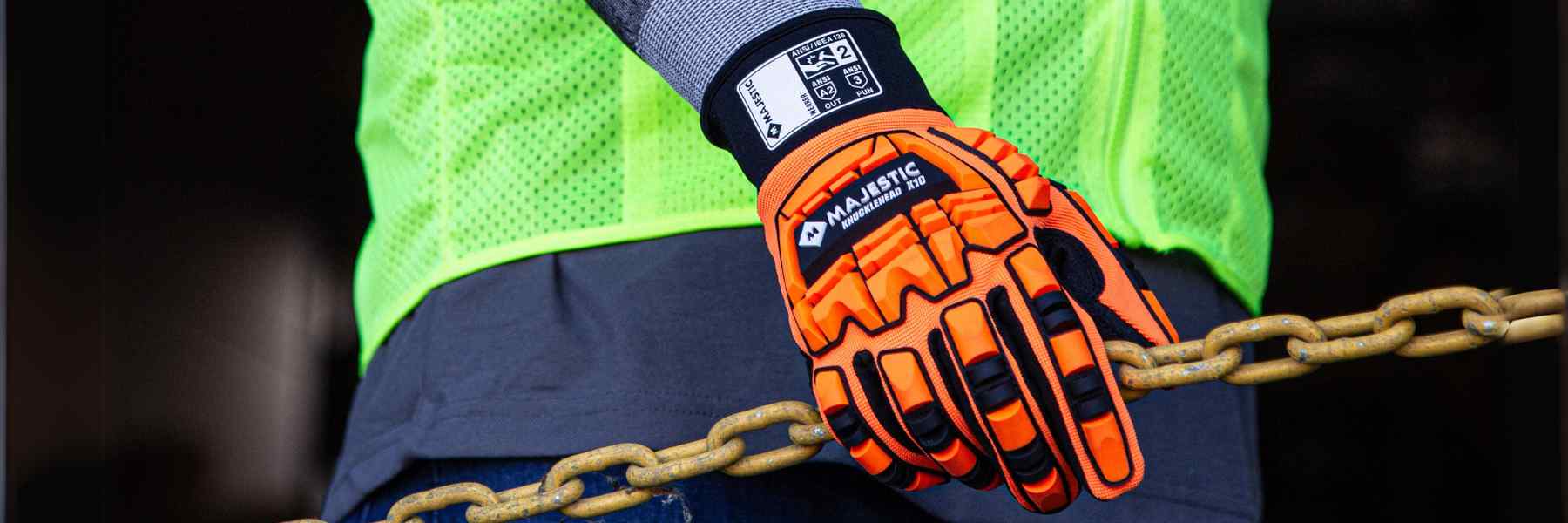 Close-up of a person’s hand wearing a high-visibility orange and black safety glove while gripping a metal chain. The glove has the word ‘MAJESTIC’ written on the cuff, and the person is wearing a neon yellow safety vest with reflective stripes partially visible in the background. The focus on the safety gear suggests an industrial or construction setting, emphasizing the importance of personal protective equipment.